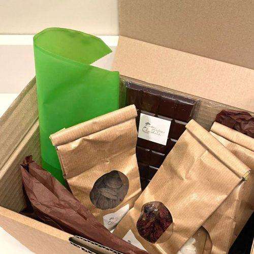 boîte en kraft ouverte avec des sachets remplis de toppings et de pastilles de chocolat, tablette de chocolat et une poche à douille verte