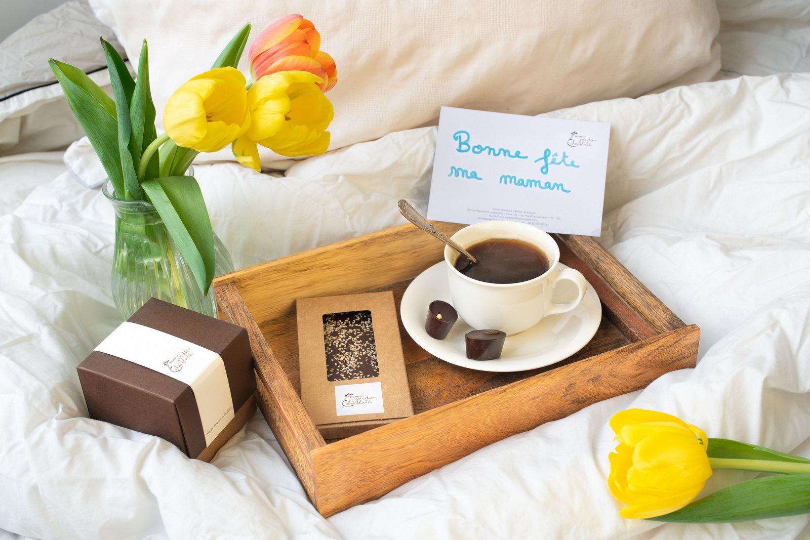 Plateau en bois contenant une tasse de café et une tablette noir sésame dans son emballage en kraft et boîte de chocolat fermée à droite